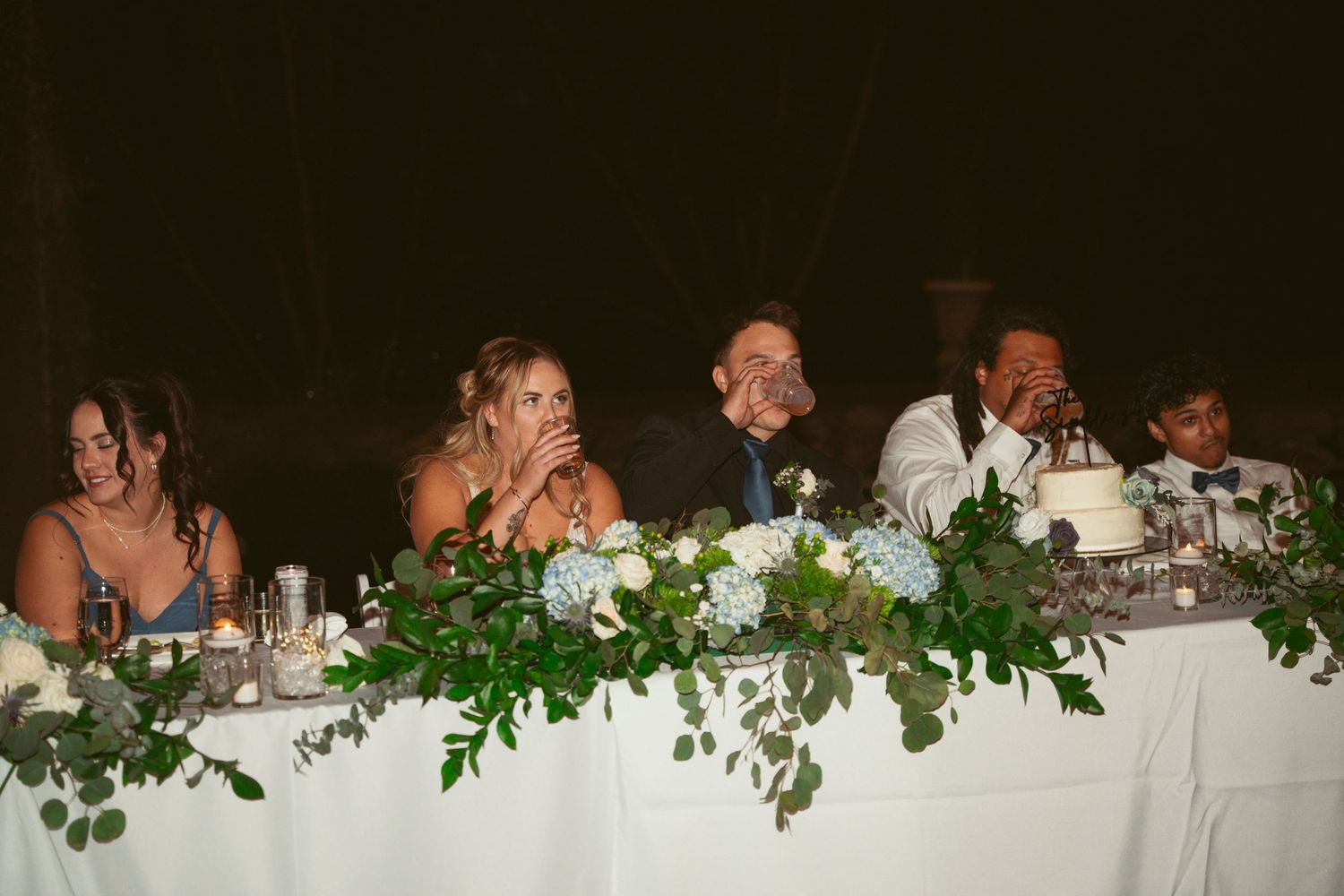Wedding party seated at a decorated head table with greenery garland and white linens.