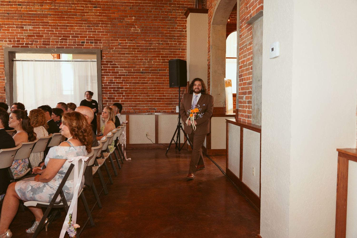 Guests seated in rows watch a wedding ceremony in an industrial brick-walled space.