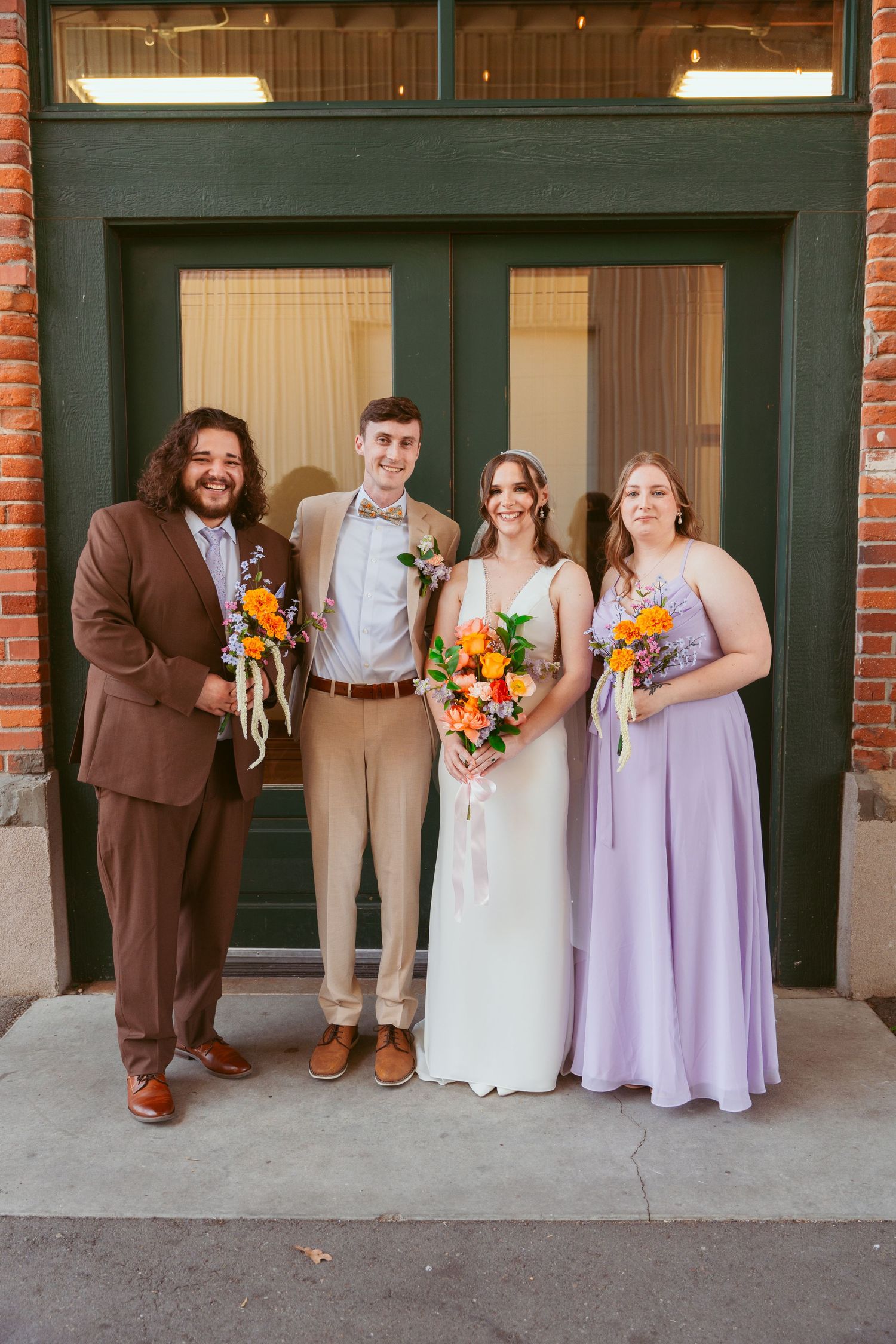 Wedding party members smile in coordinated attire of brown suits, white and lavender dresses.