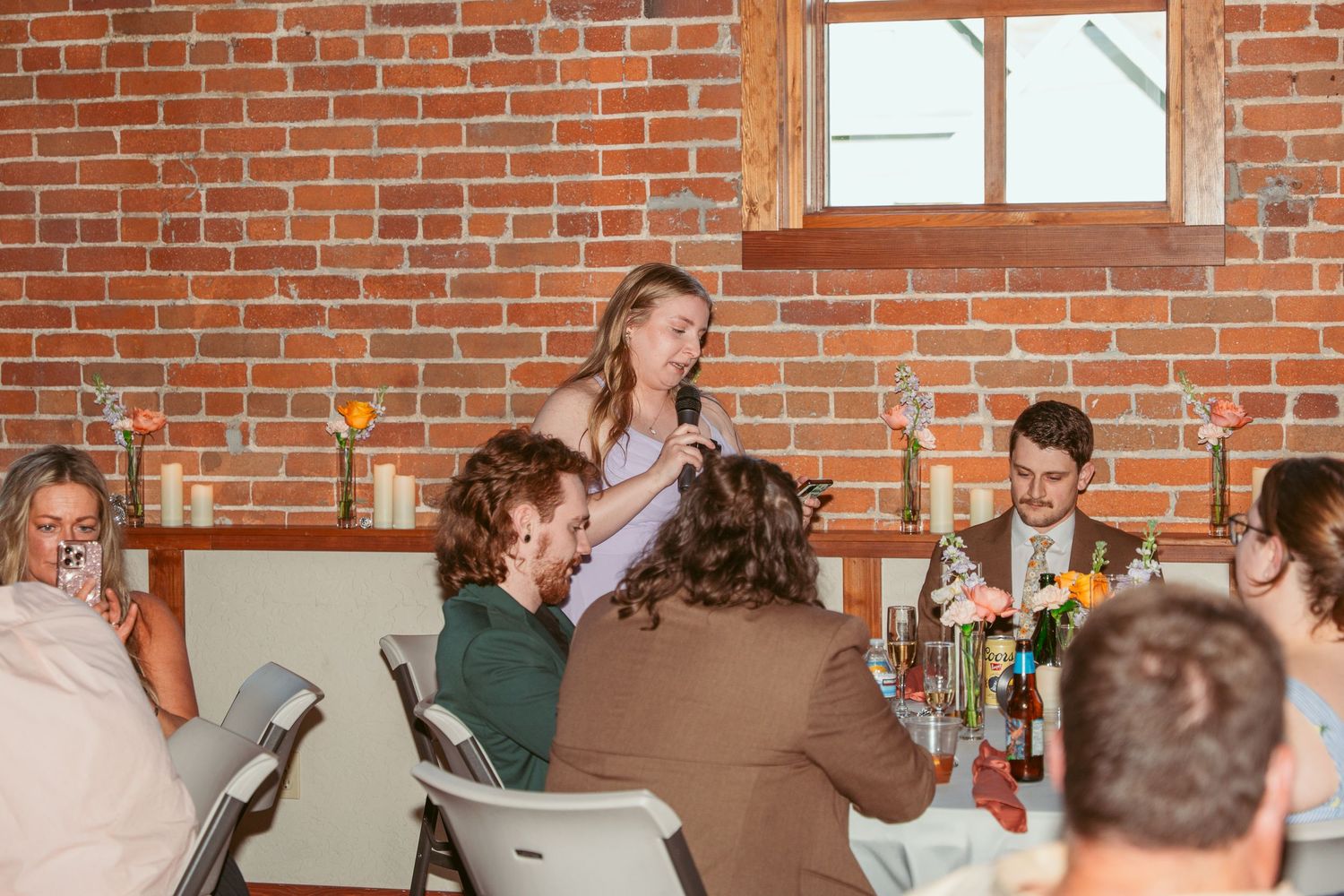 Wedding reception guests socializing at tables in a rustic brick venue with natural lighting.