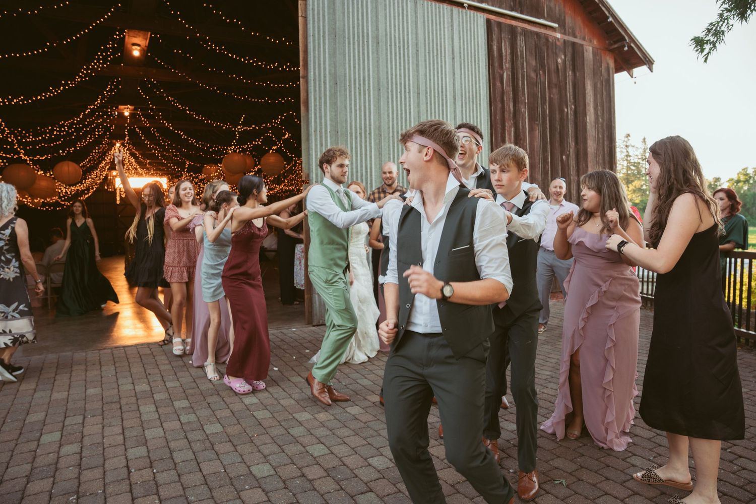 Wedding party dances in synchronized formation on brick patio under twinkling lights.