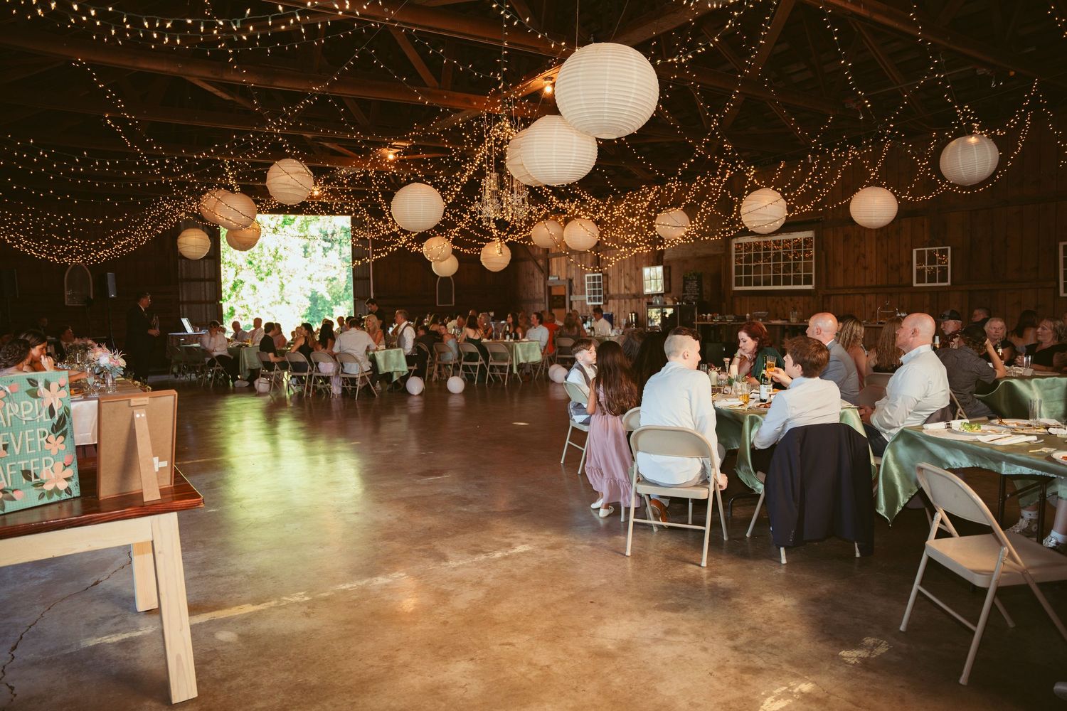 Wedding reception venue with string lights and paper lanterns illuminating tables of guests in a rustic barn.