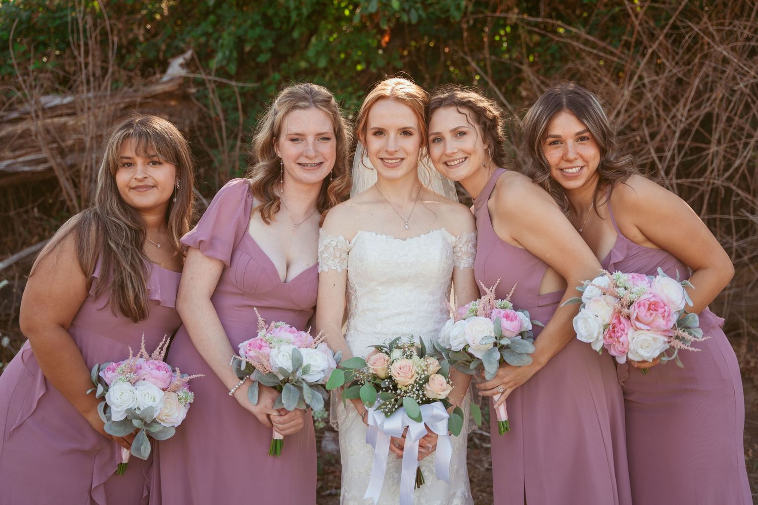 Wedding party members pose together with pink and white bouquets outdoors.