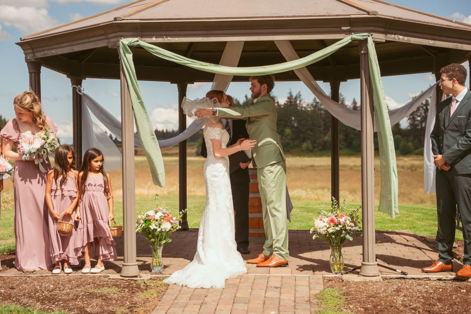 Wide angle shot of a wedding ceremony under a covered pavilion with guests seated on rustic wooden chairs.