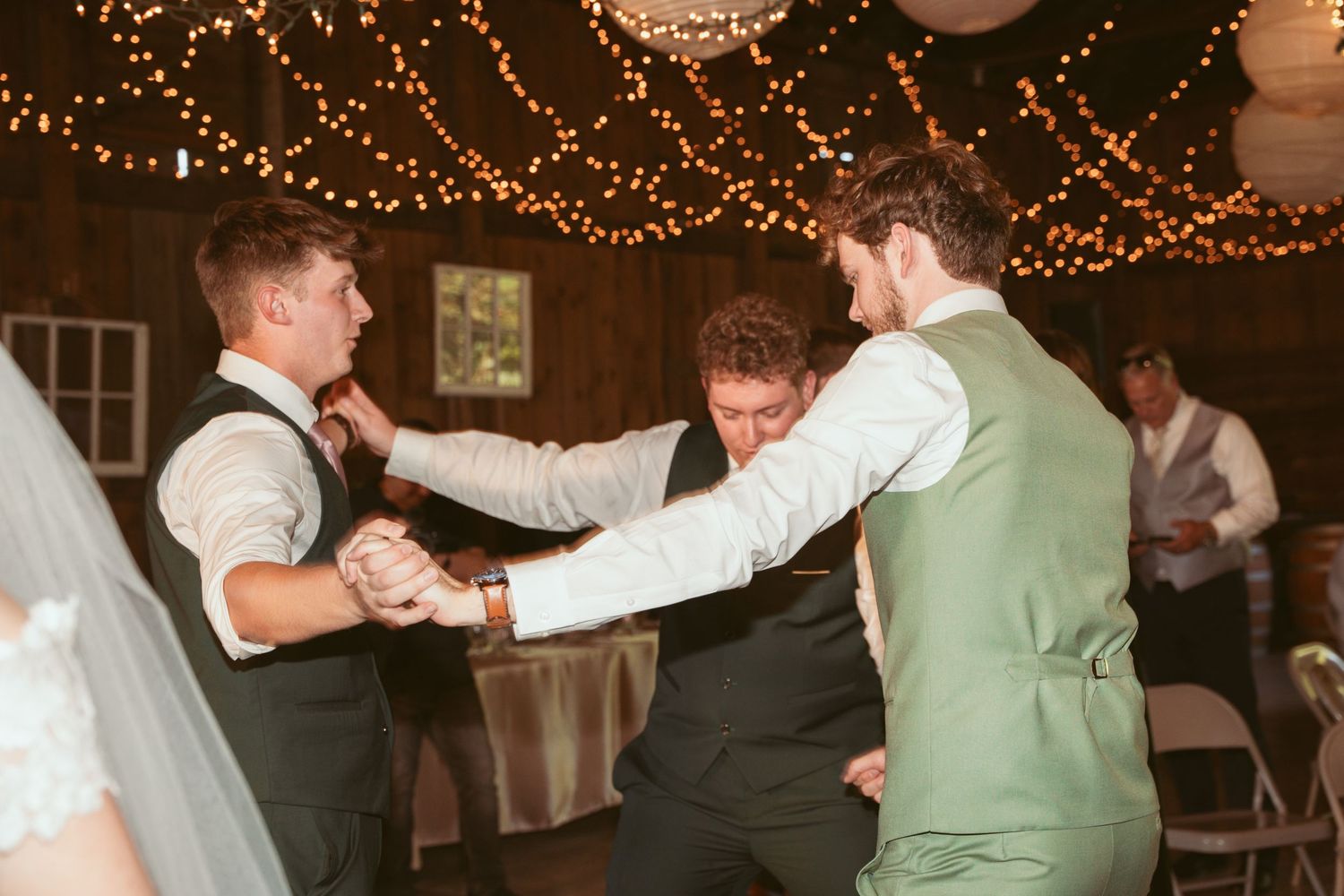 Guests dancing and celebrating at a barn reception with twinkle lights and paper lanterns overhead.