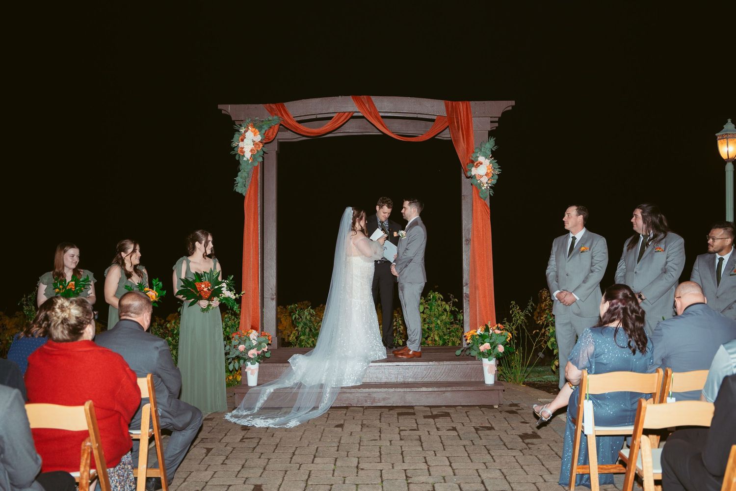 Wedding ceremony space with wooden chairs and red-draped arch illuminated by string lights.