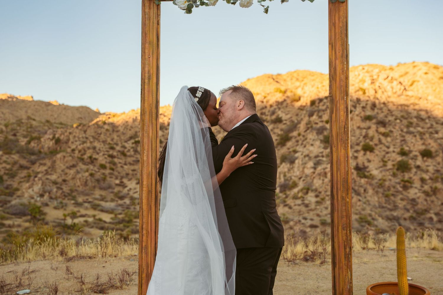 A wedding couple poses under a rustic wooden arch decorated with greenery in a desert landscape.