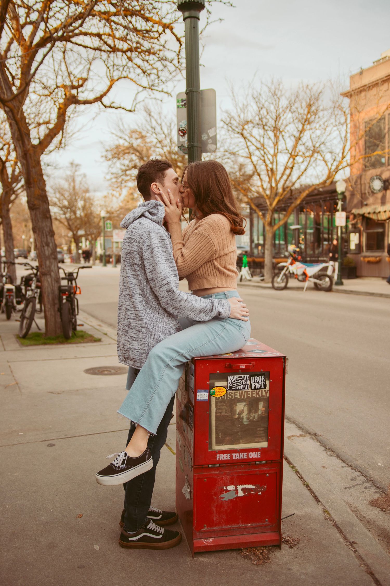 A couple shares a romantic moment on a red newspaper box on a tree-lined street.