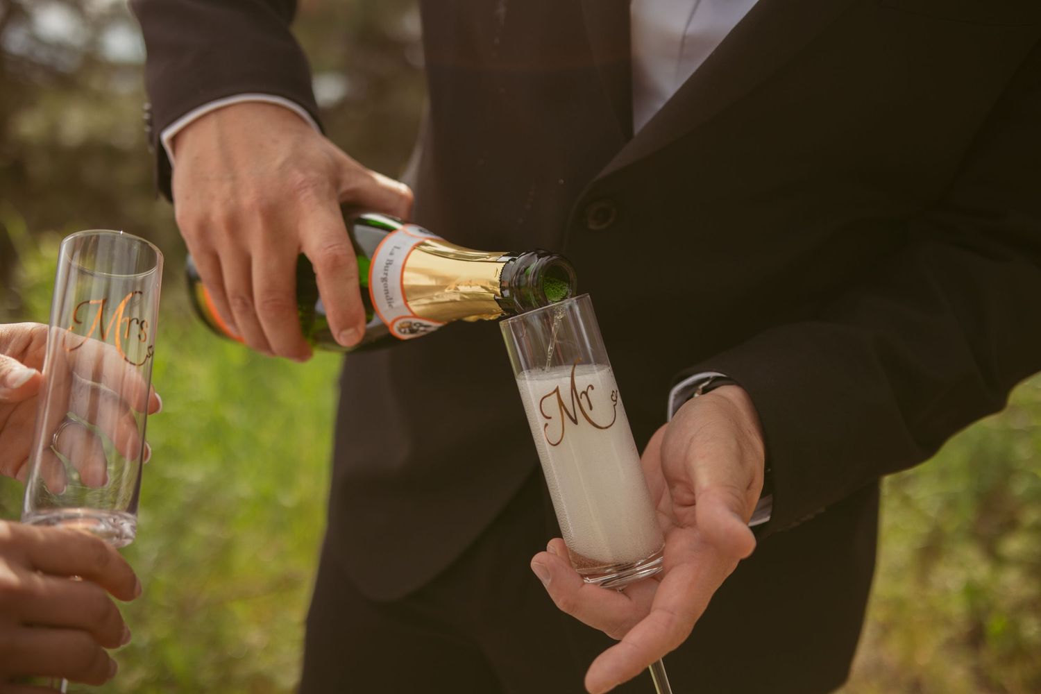 Close-up of hands pouring champagne into wedding glasses during an outdoor celebration.