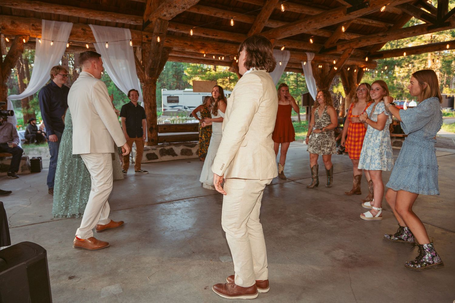 Guests line dancing at a rustic outdoor wedding reception under wooden beams and lights.