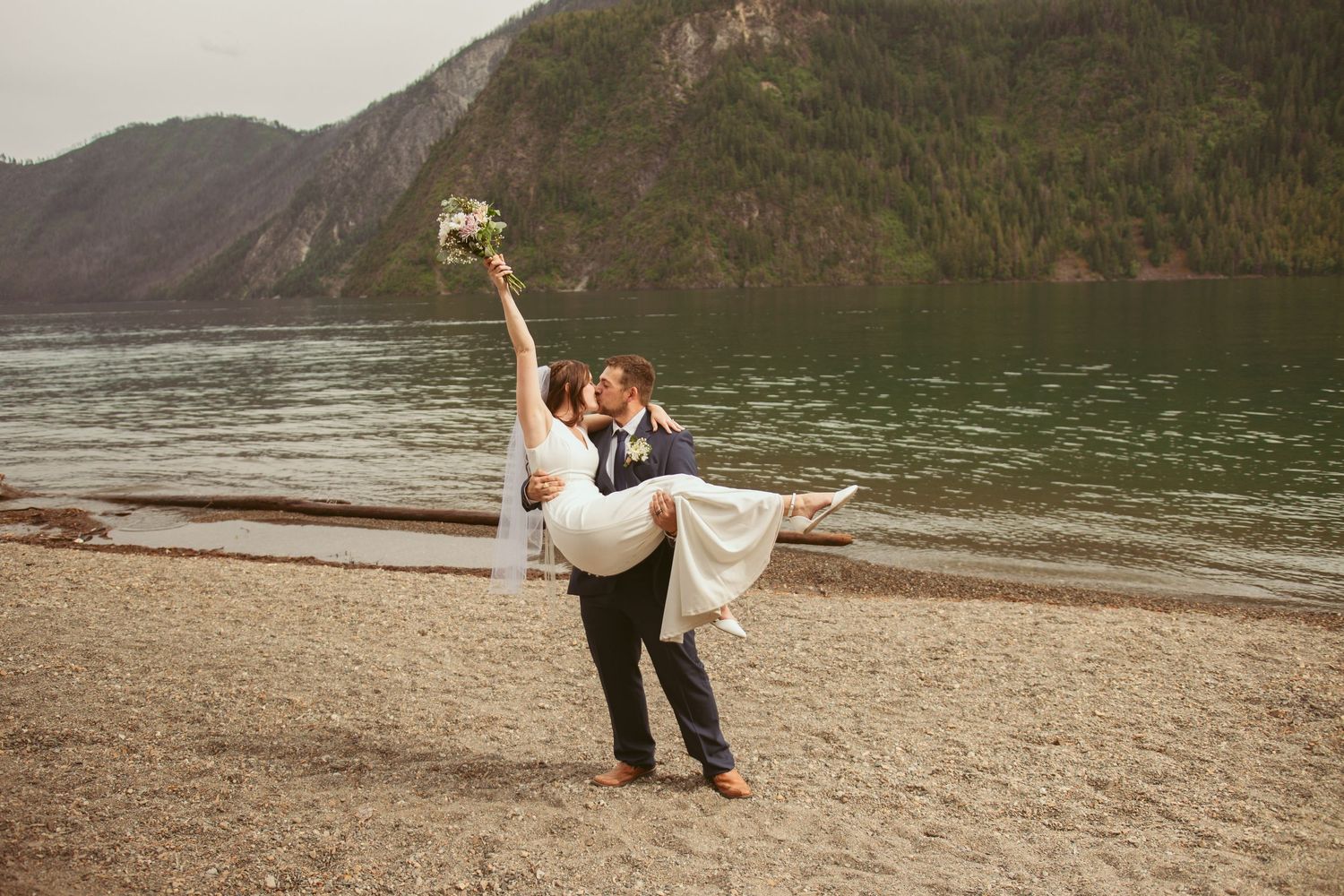 Romantic couple dances and poses along a scenic lakeside beach with mountains in the background.