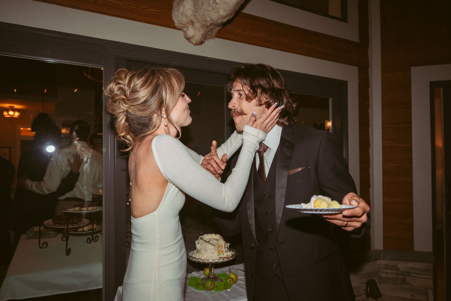 A couple shares wedding cake in a romantic indoor setting with ambient lighting.