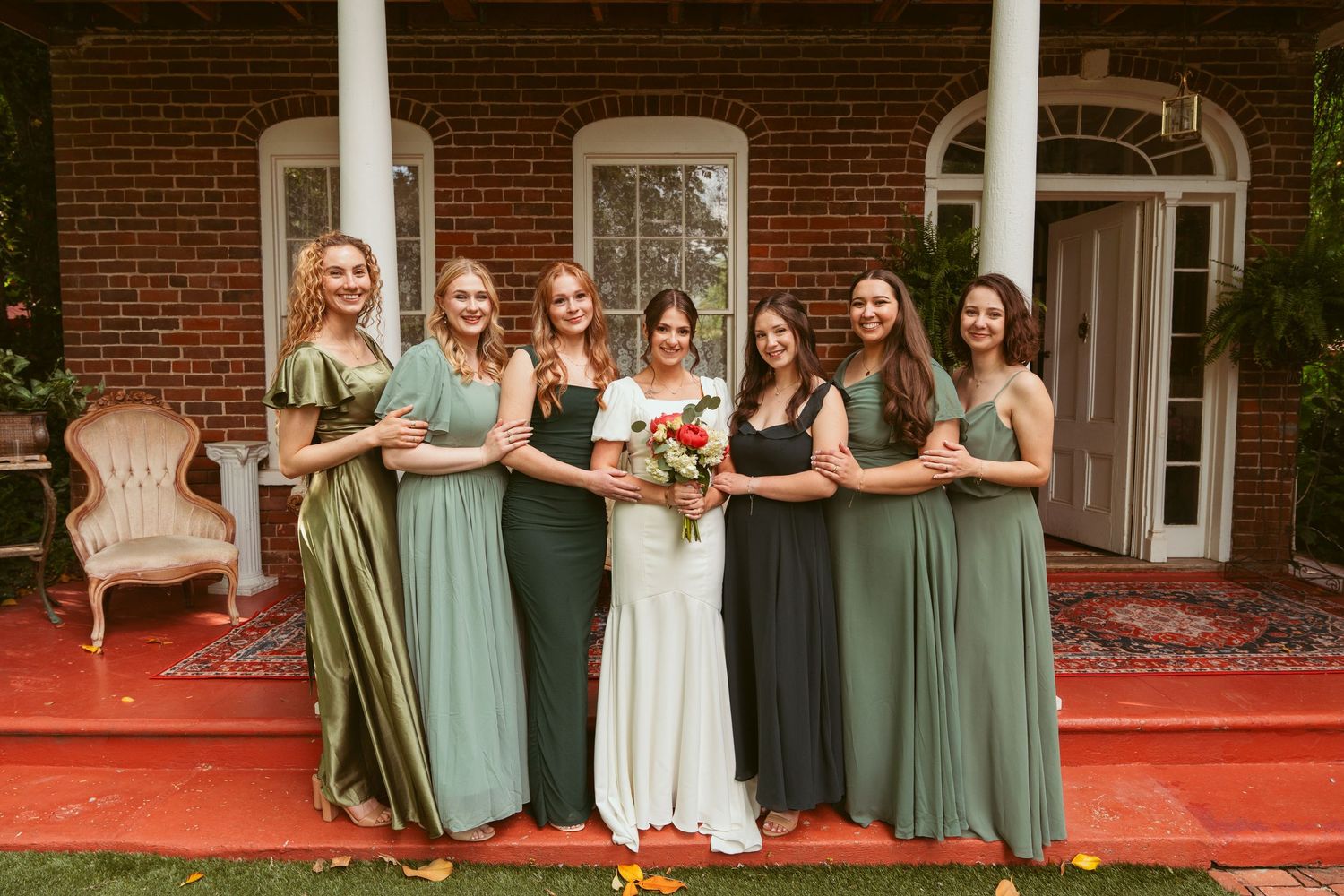 Bridesmaids in various shades of green dresses pose on a vintage porch with red carpet.
