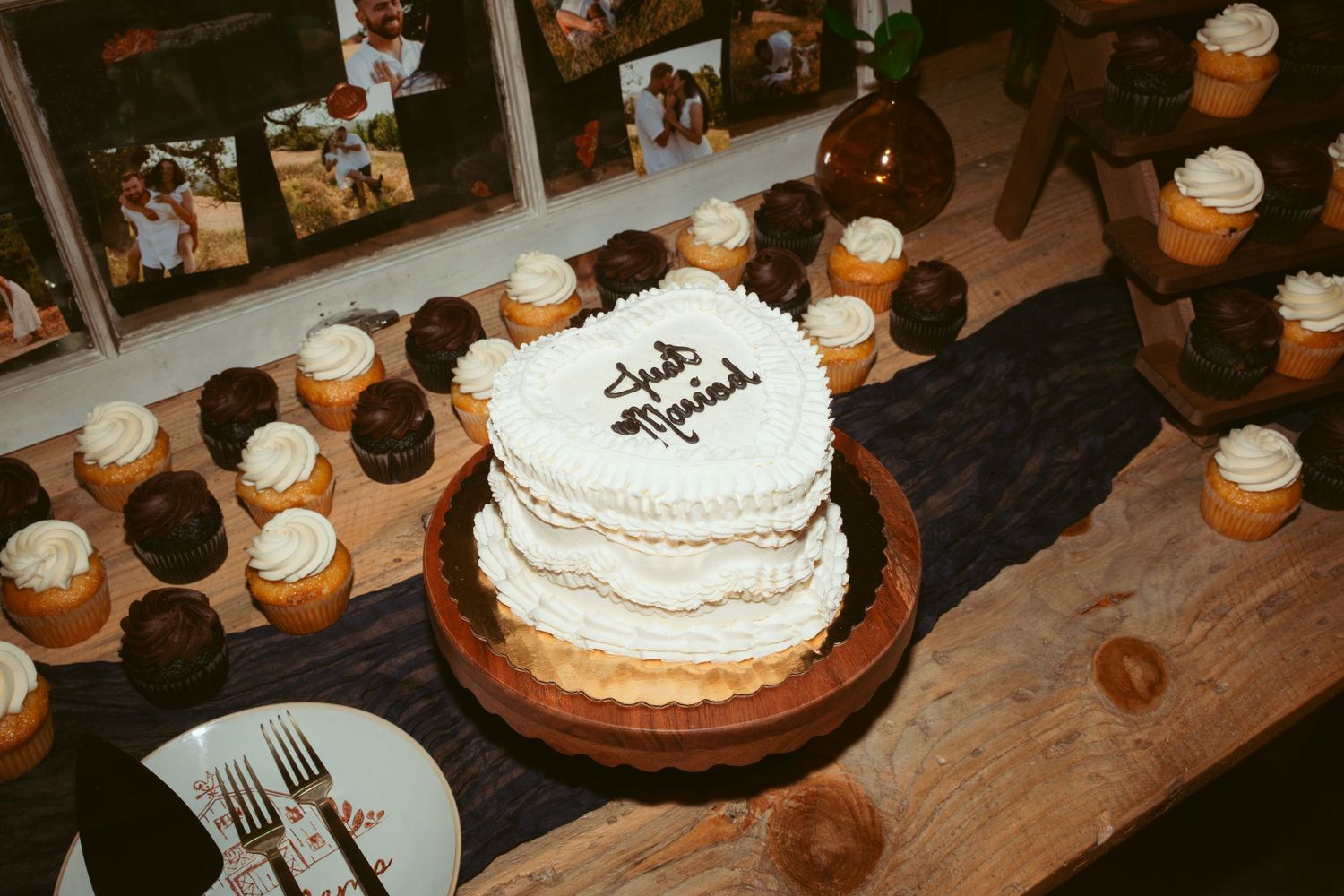 White tiered wedding cake surrounded by cupcakes on wooden display table.
