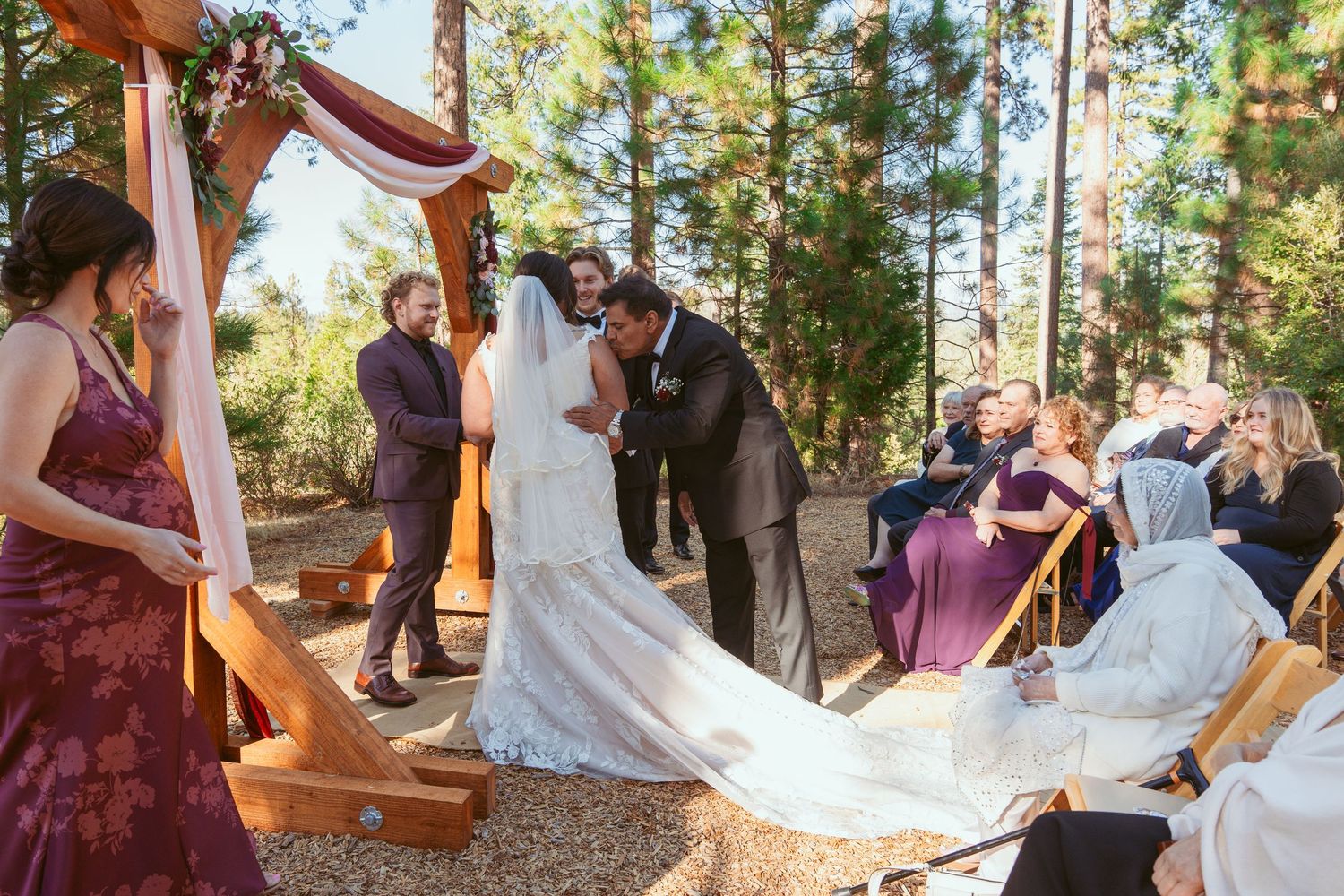 An outdoor forest wedding ceremony with guests seated on wooden chairs among tall evergreen trees.