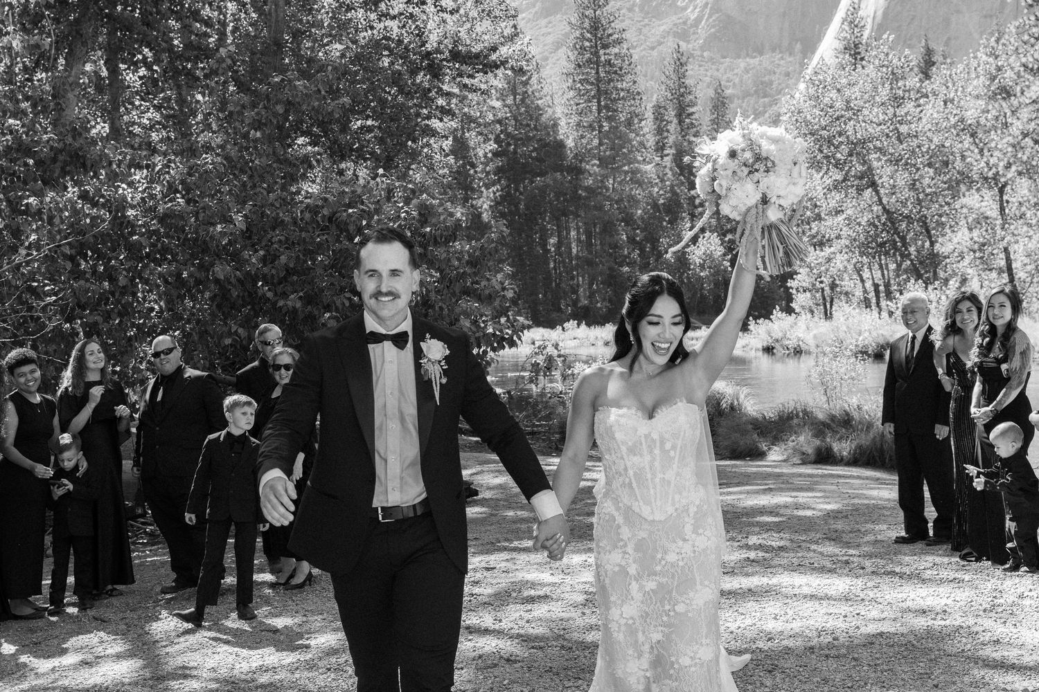 Joyful newlyweds walk hand-in-hand after outdoor wedding ceremony in Yosemite, bride raising bouquet in black and white photo.