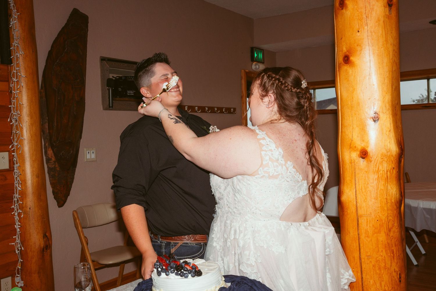A couple shares a messy cake-smashing moment at their wedding reception next to a wooden pillar.
