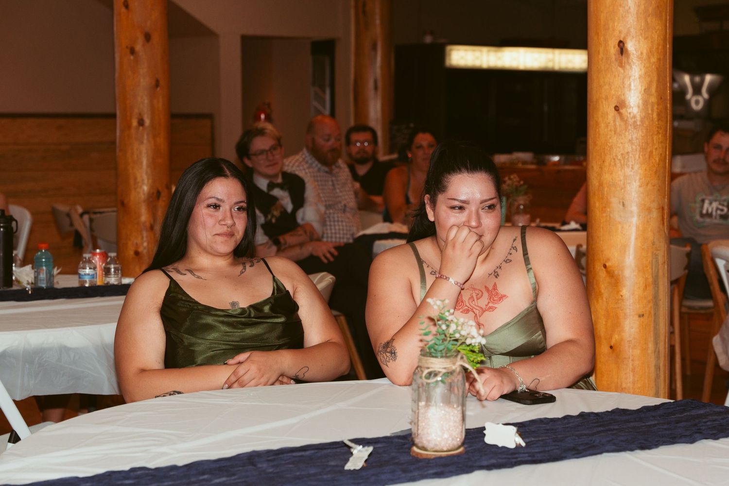 Wedding reception guests seated at long tables with wooden log pillars in the background.
