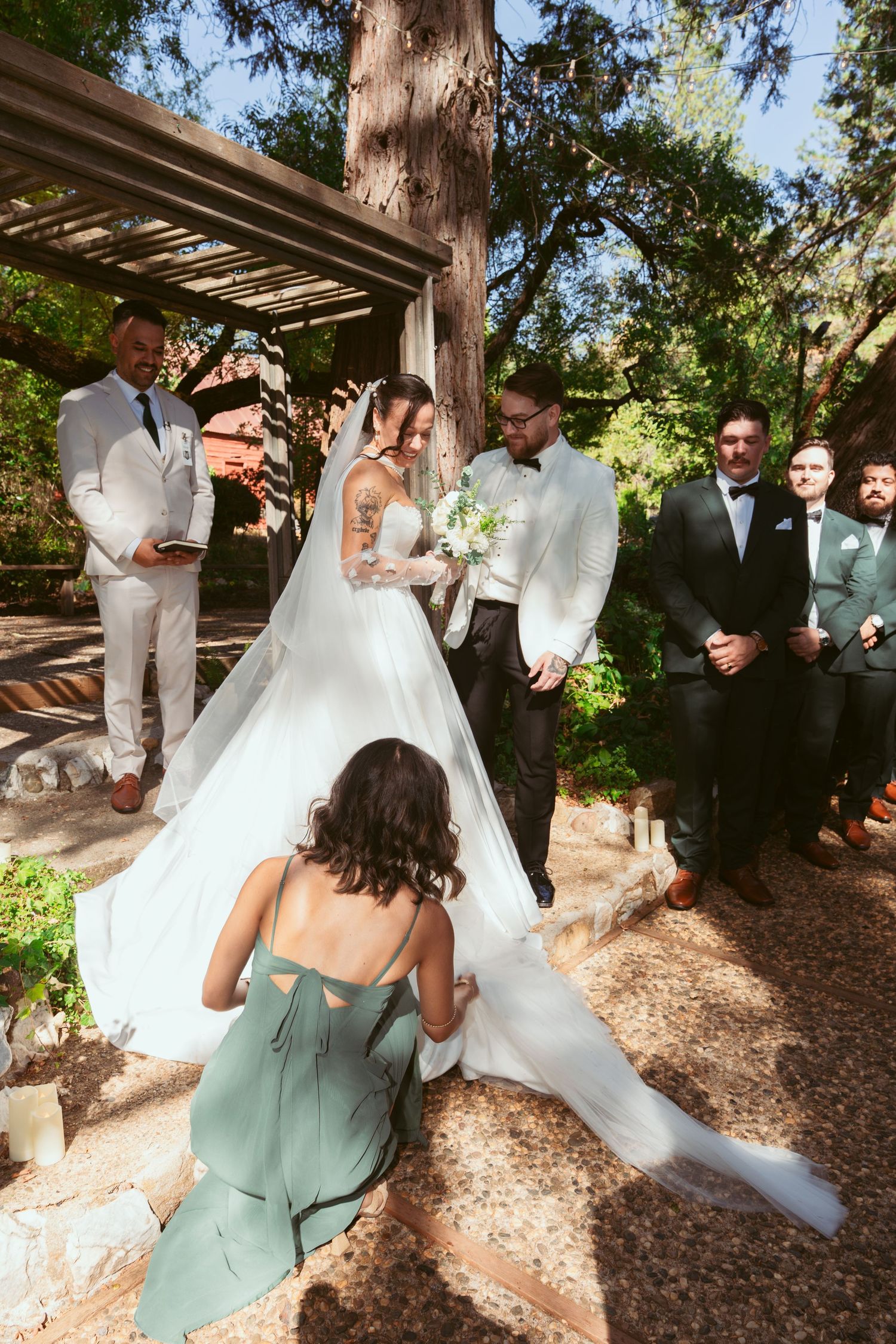 Wedding party helps adjust a flowing white wedding dress train in a shaded garden path.