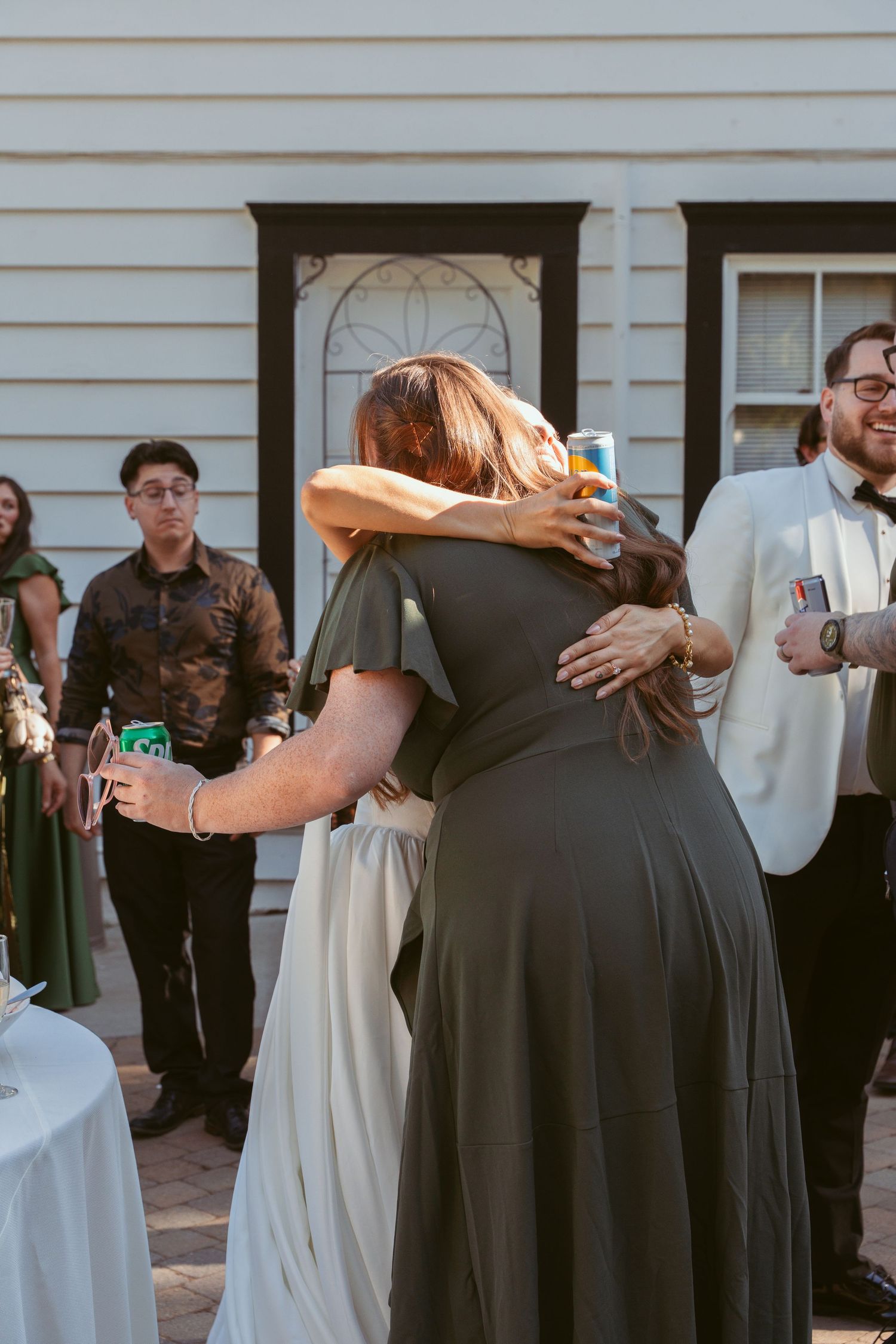 Wedding party members embrace in a group hug outside a white building.