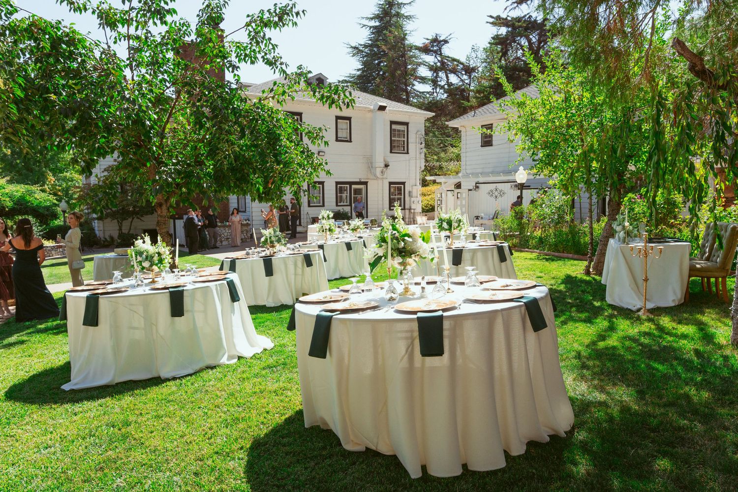 Long banquet tables with white tablecloths set up outdoors in a lush garden setting for an elegant event.