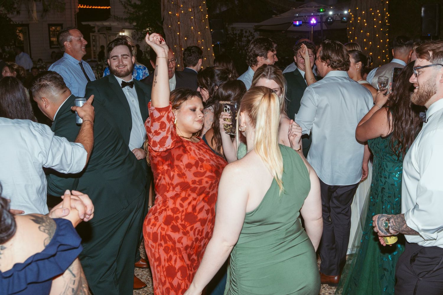 Lively outdoor wedding reception with guests dancing under string lights at dusk.