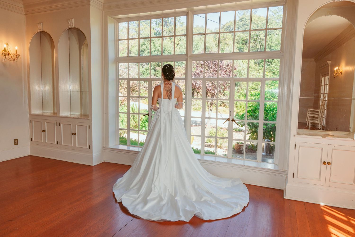 A bride in an elegant white gown with a long train stands before a large window in a sunlit room.