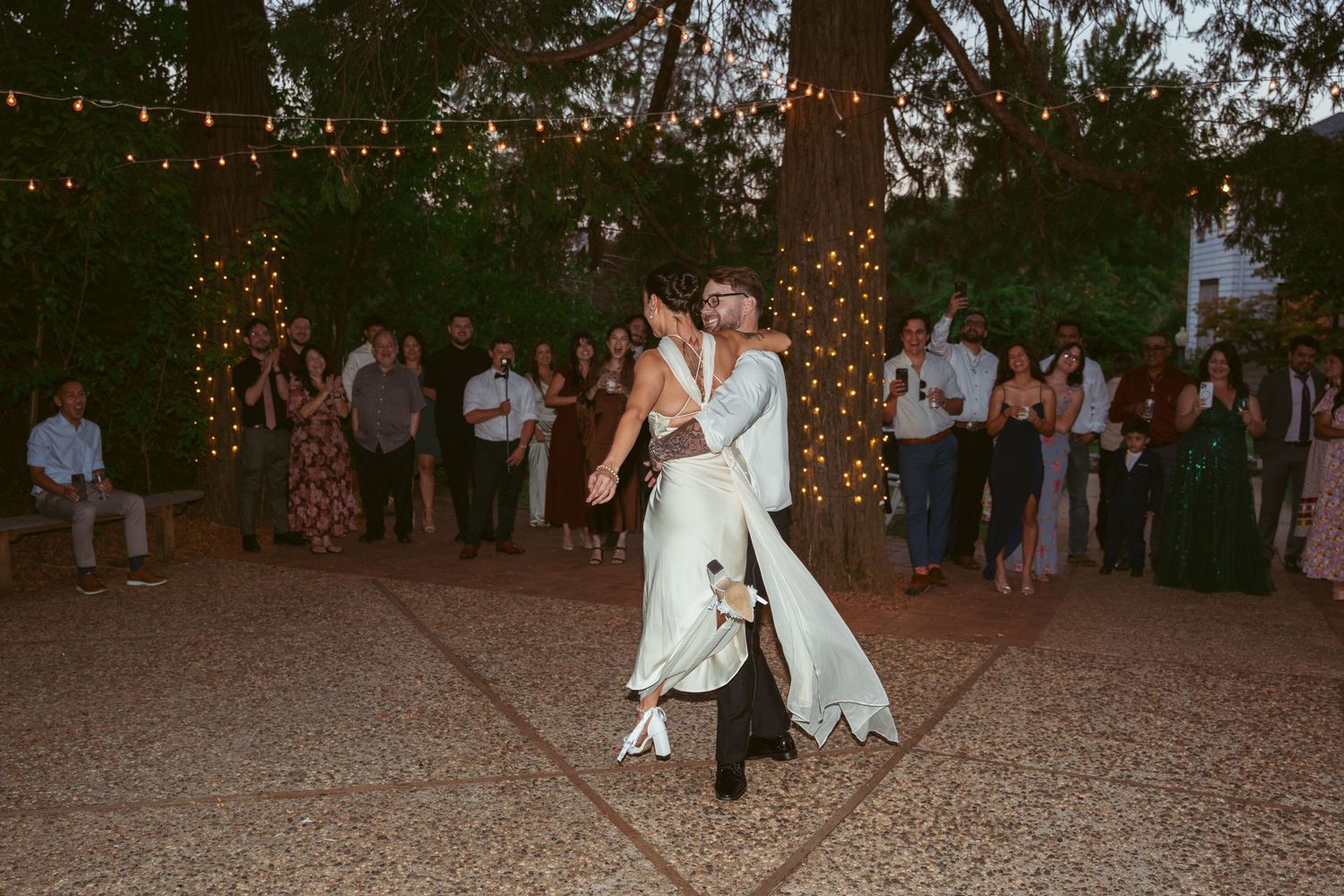 A couple shares their first dance under twinkling lights strung between trees at an outdoor wedding reception.
