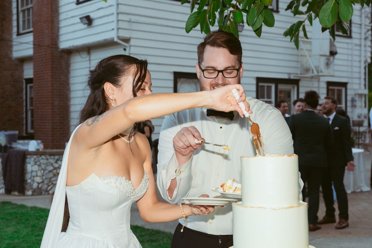 Newlyweds playfully feeding each other wedding cake at outdoor celebration.