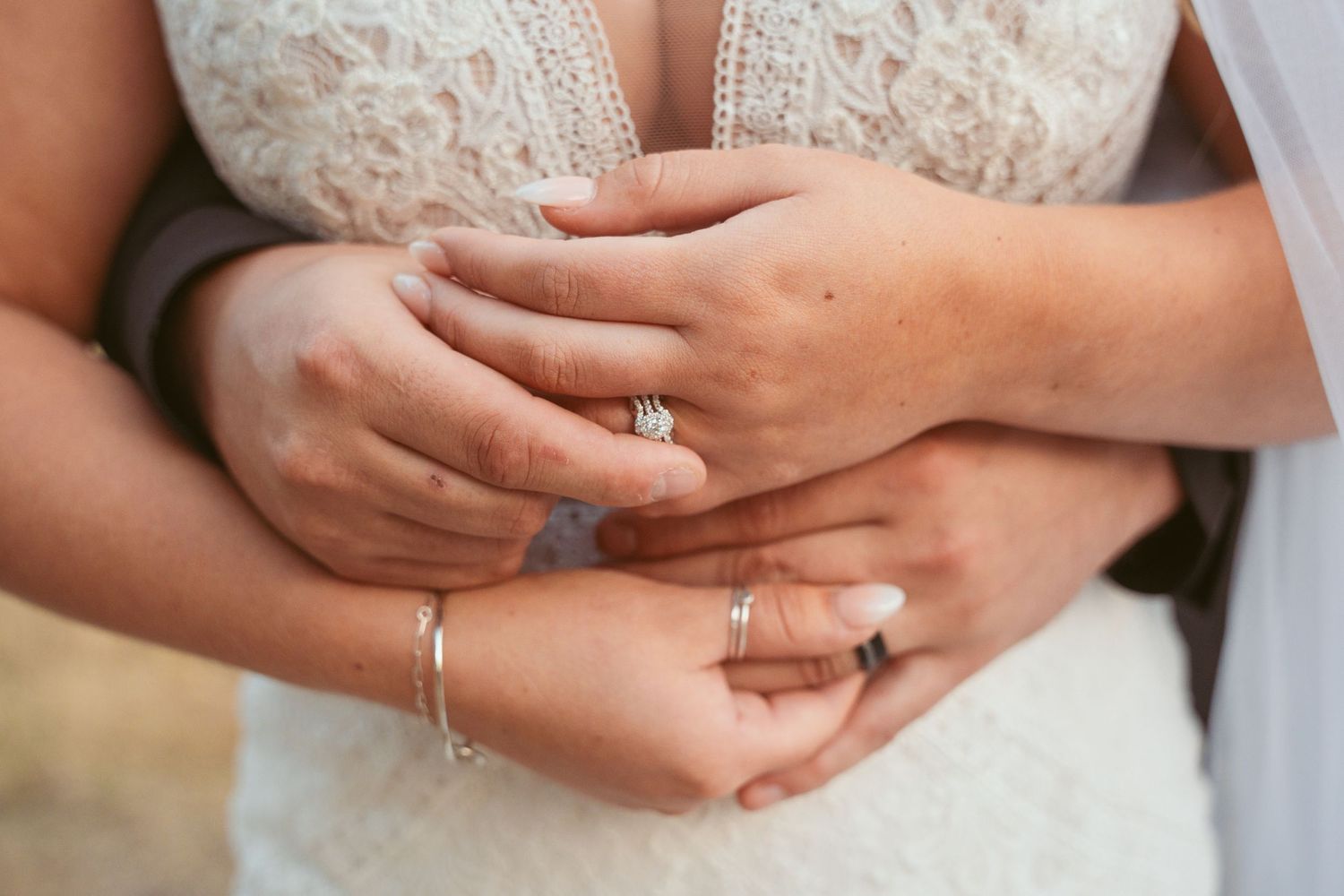 Close-up of hands with wedding rings intertwined against white lace dress.
