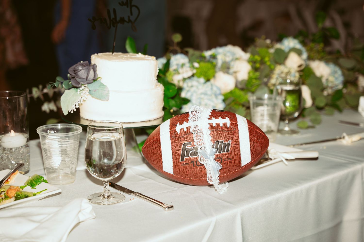 Wedding reception table decorated with white linens, a small white cake, and a football centerpiece on display.