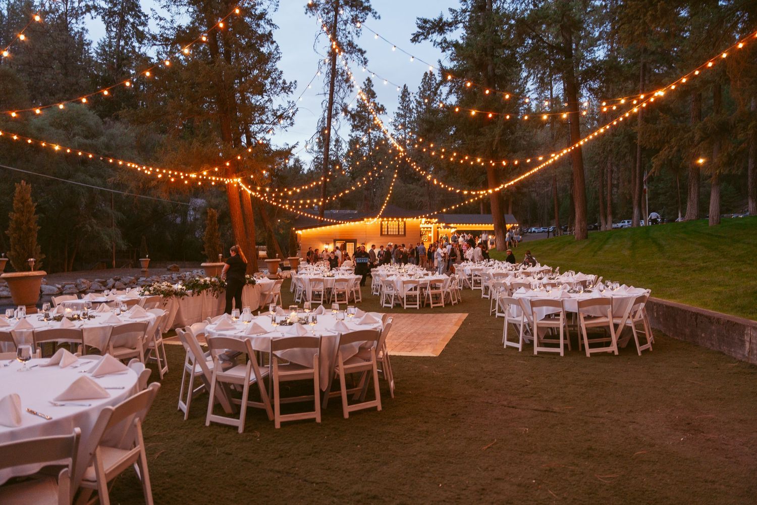 Outdoor wedding reception venue with string lights and white tables set up in a forest clearing at dusk.
