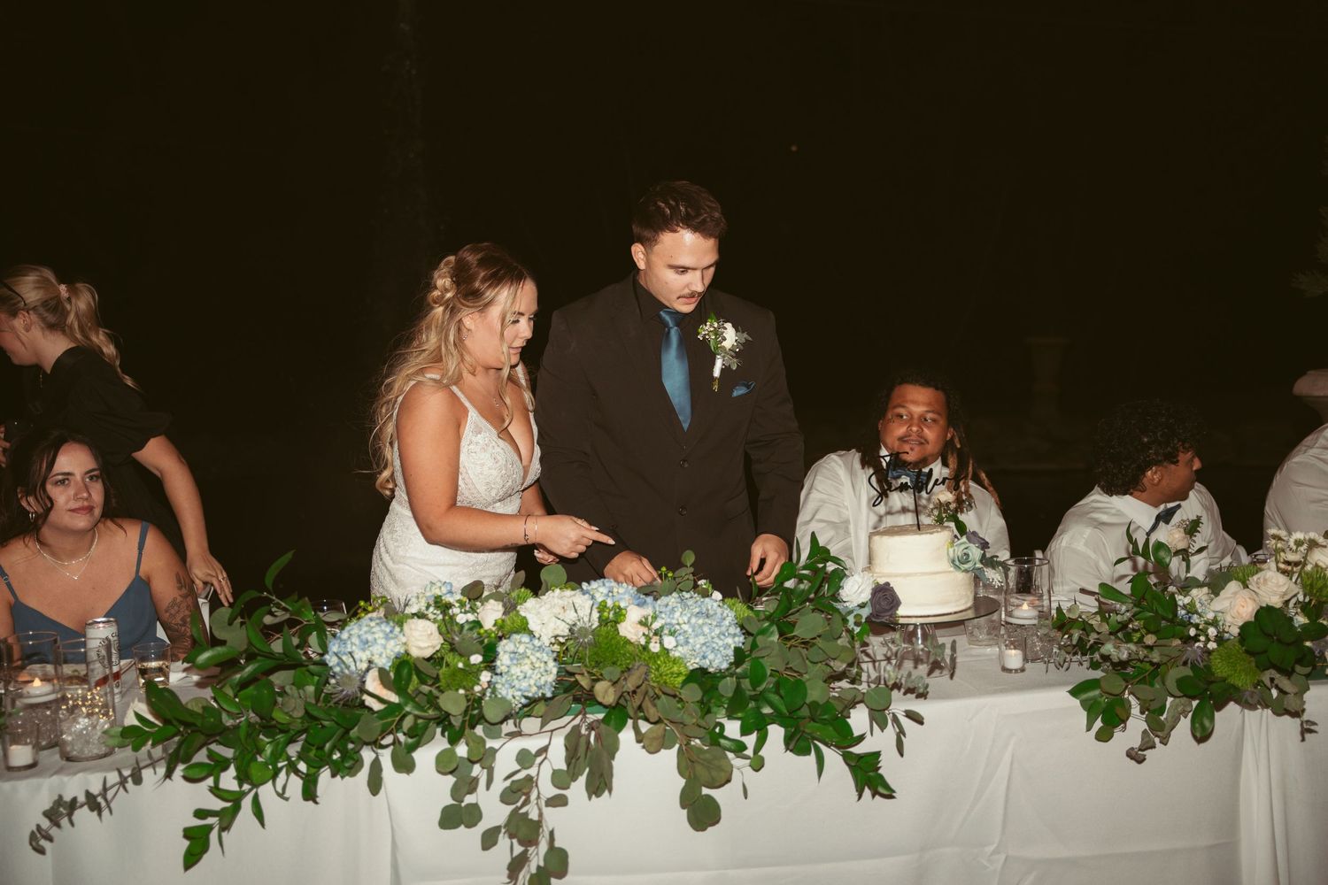 Wedding party seated at decorated head table with greenery garland and white linens.