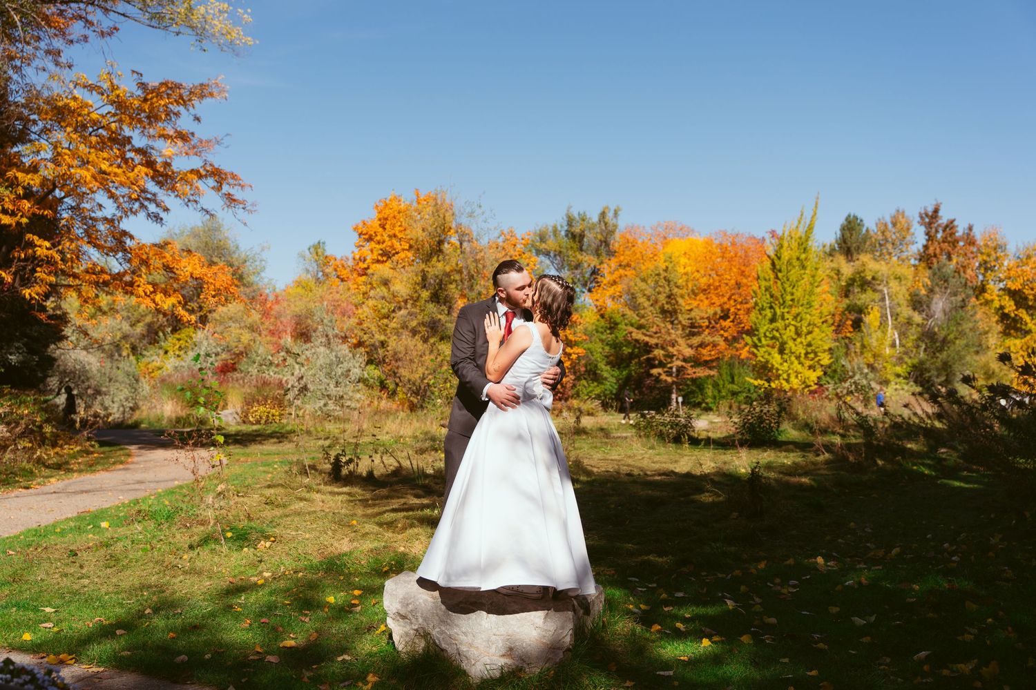 A wedding couple poses on a rock with beautiful autumn foliage in the background.