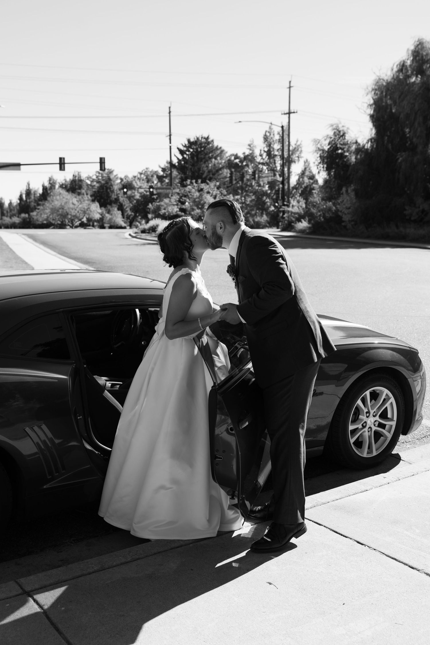 A couple in wedding attire share a tender moment by a black car on a sunny day.