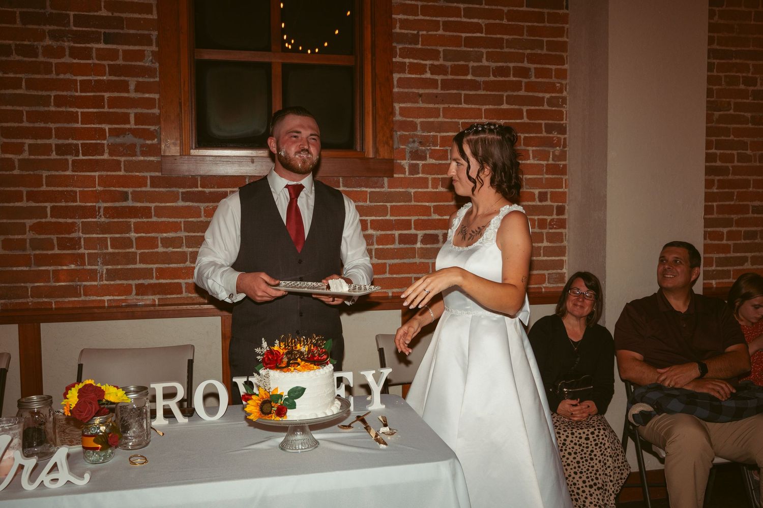 Couple laughing while cutting their wedding cake together at reception with exposed brick wall behind them.