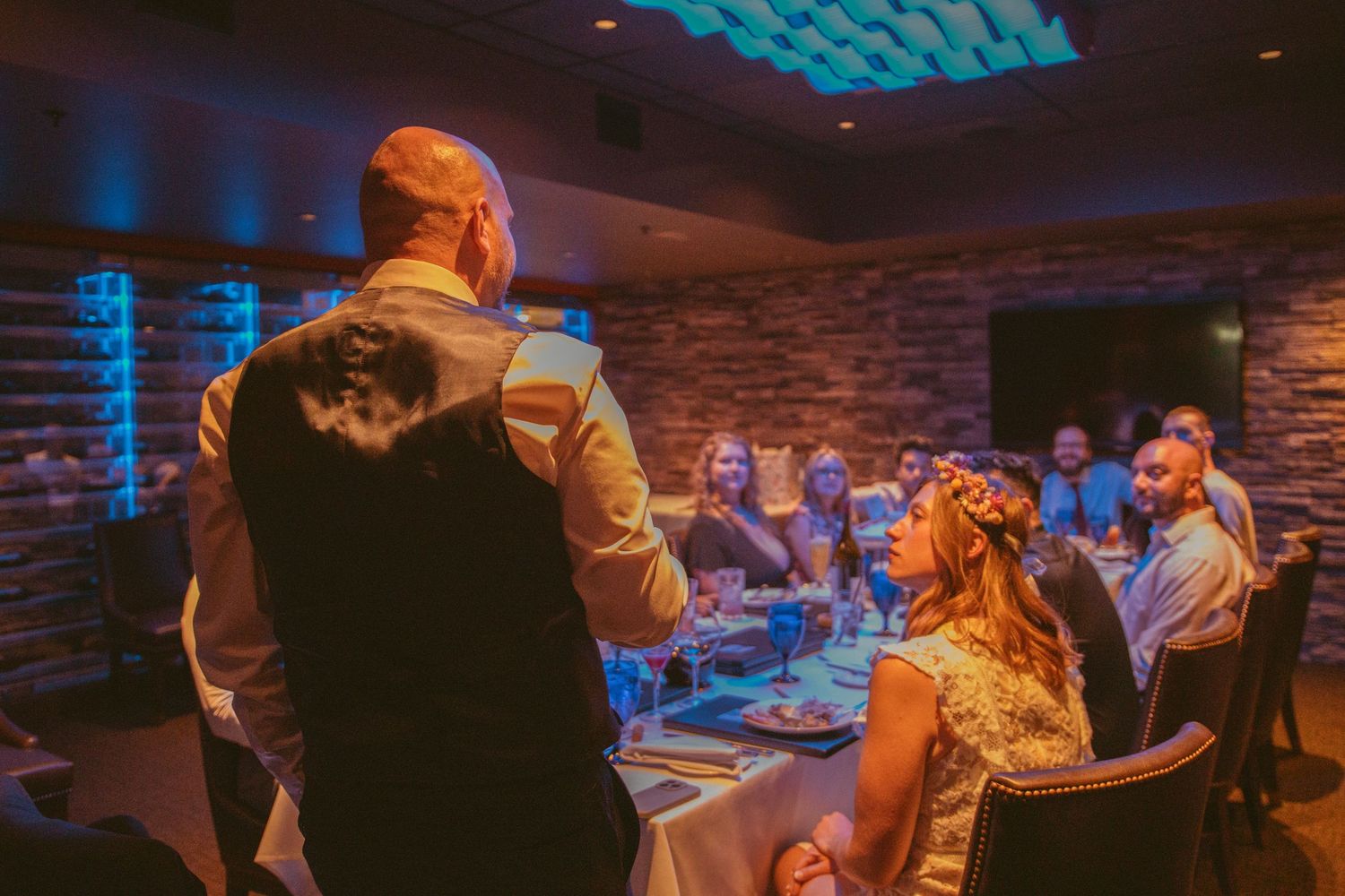 Wedding guests listen attentively during a reception speech in a rustic venue with mood lighting.