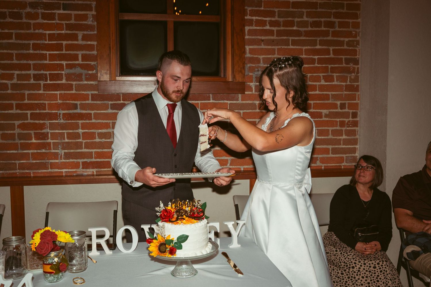 Wedding cake cutting ceremony with sunflowers and decorations against exposed brick wall.