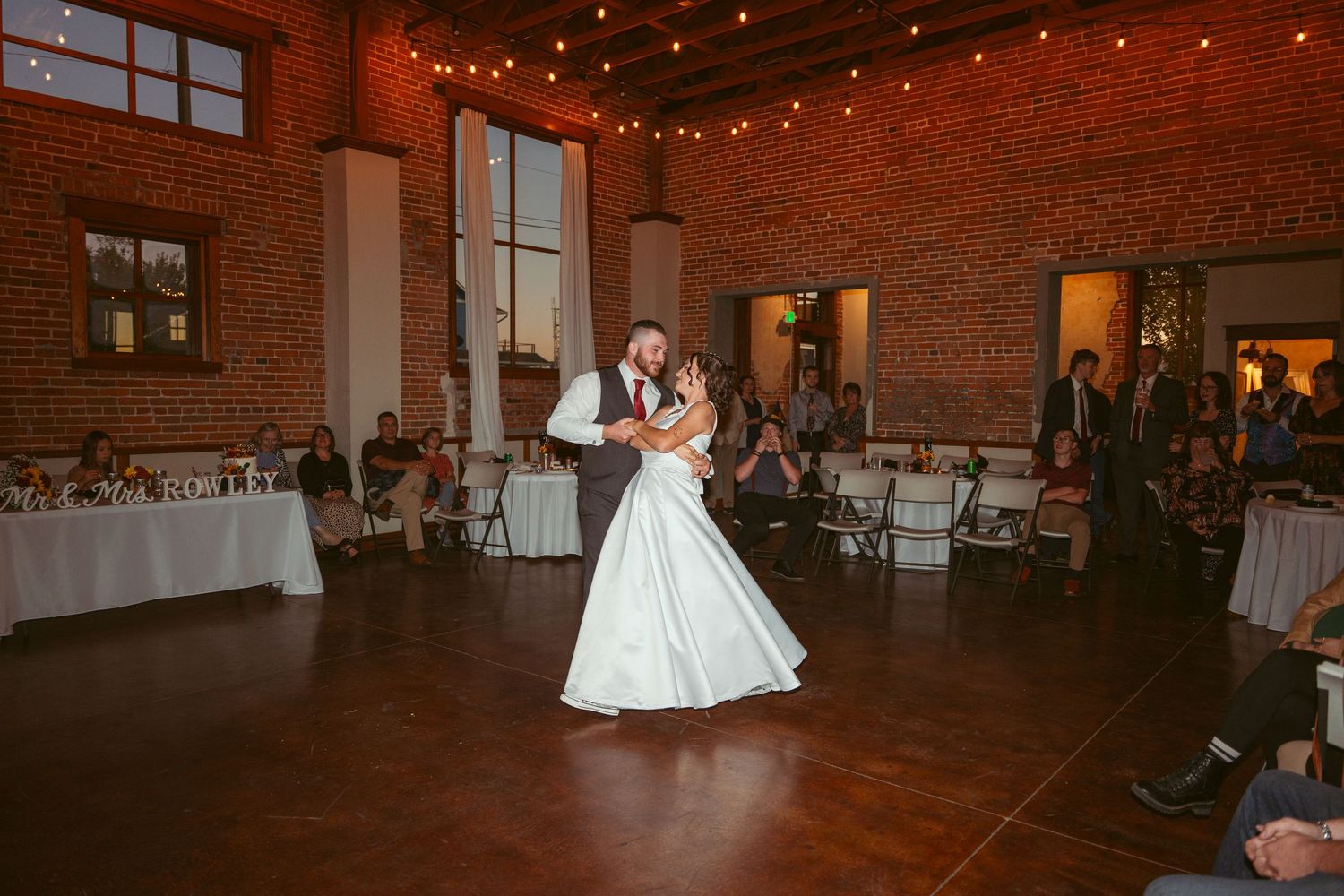 A romantic dance sequence unfolds in a warmly lit brick venue with string lights and white table settings.