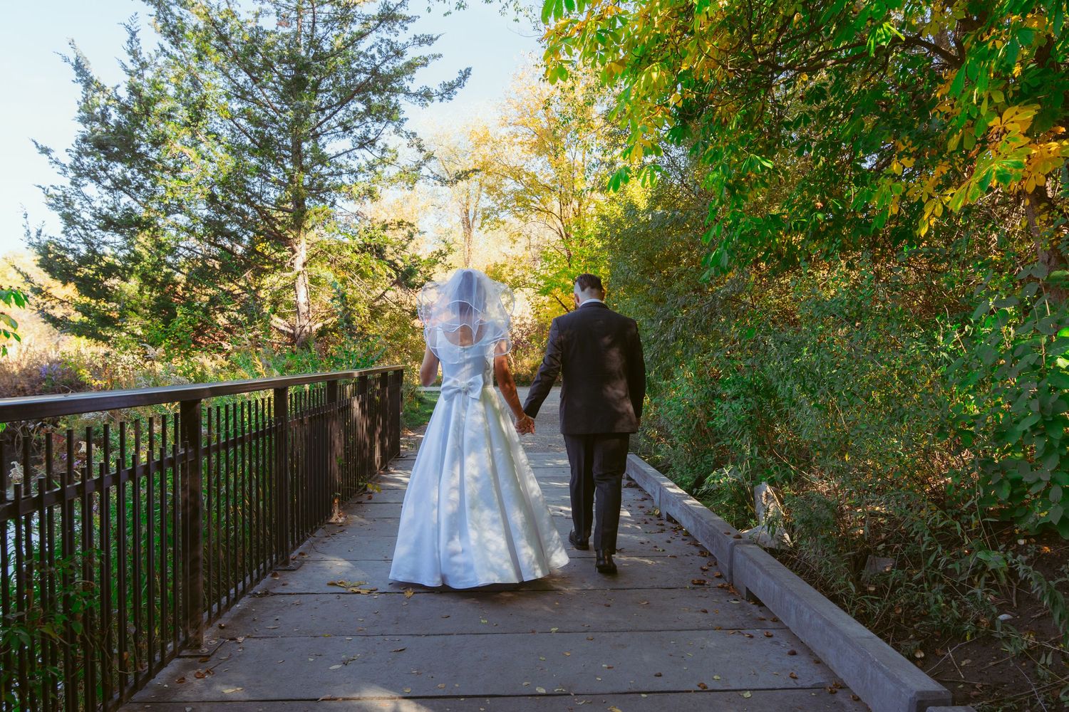 Bride and groom walking along tree-lined pathway during autumn wedding.