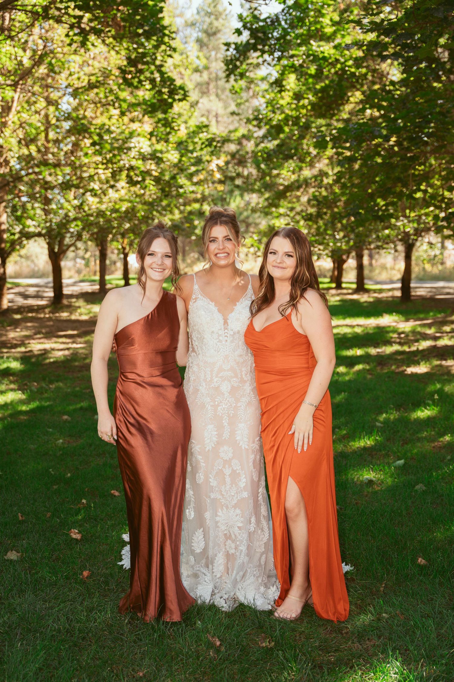 Three women pose outdoors in a wooded setting wearing elegant dresses in rust orange and white colors.