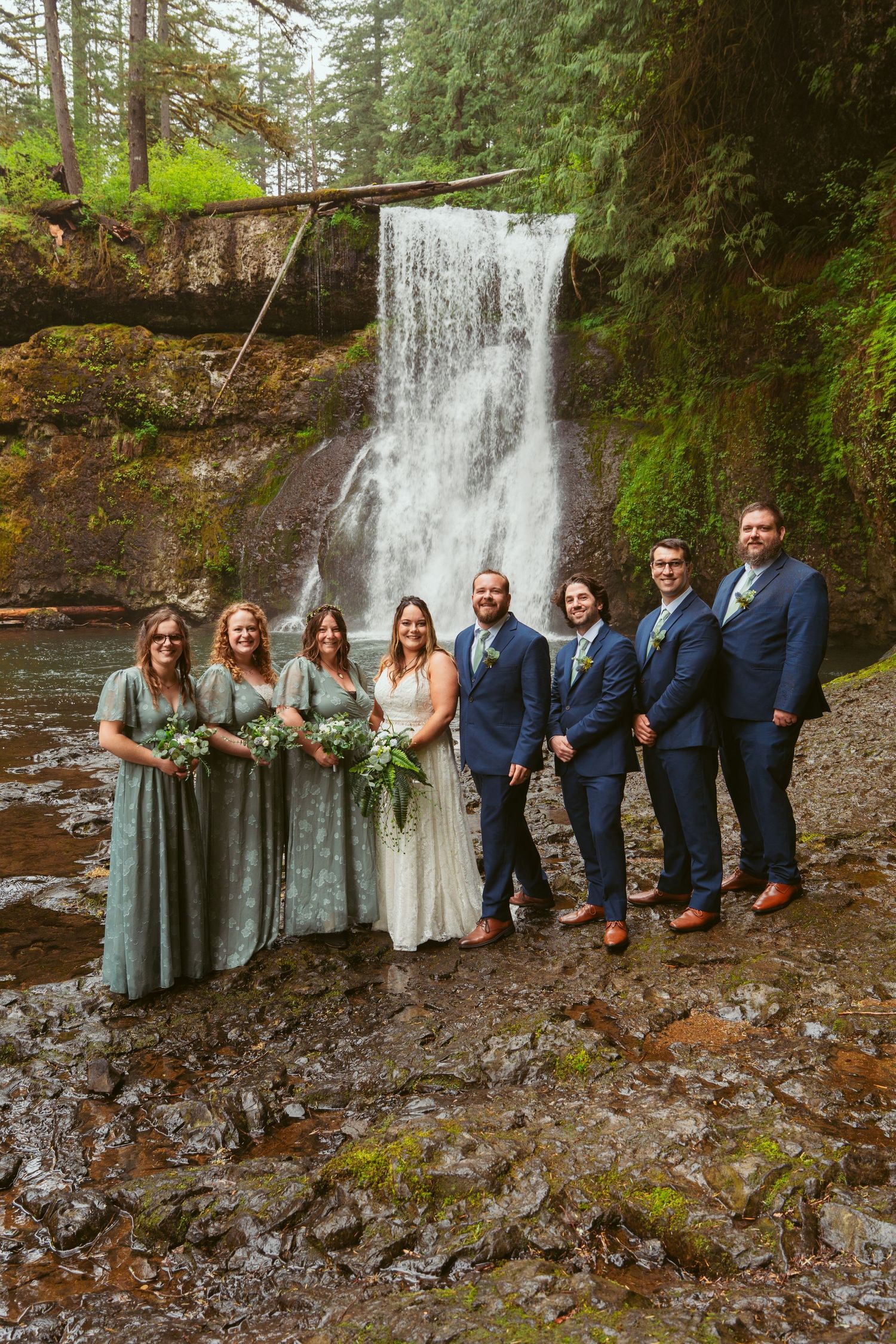 Wedding party members in navy suits and sage dresses arranged casually by forest waterfall.