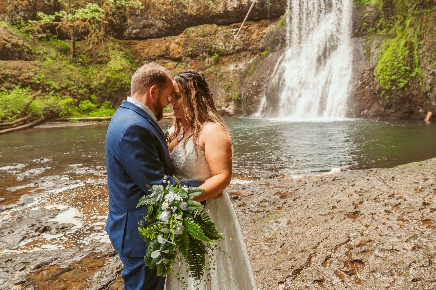 Newlyweds embrace by cascading waterfall during outdoor photo session.
