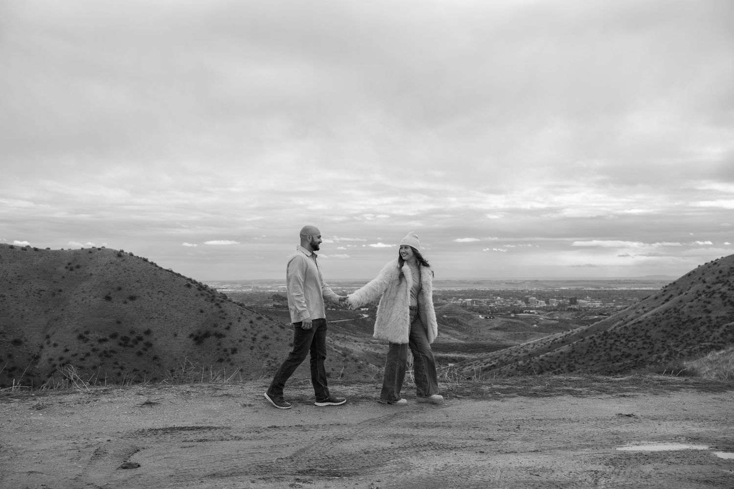 A couple holds hands while standing on a hilltop against a dramatic cloudy sky in black and white.
