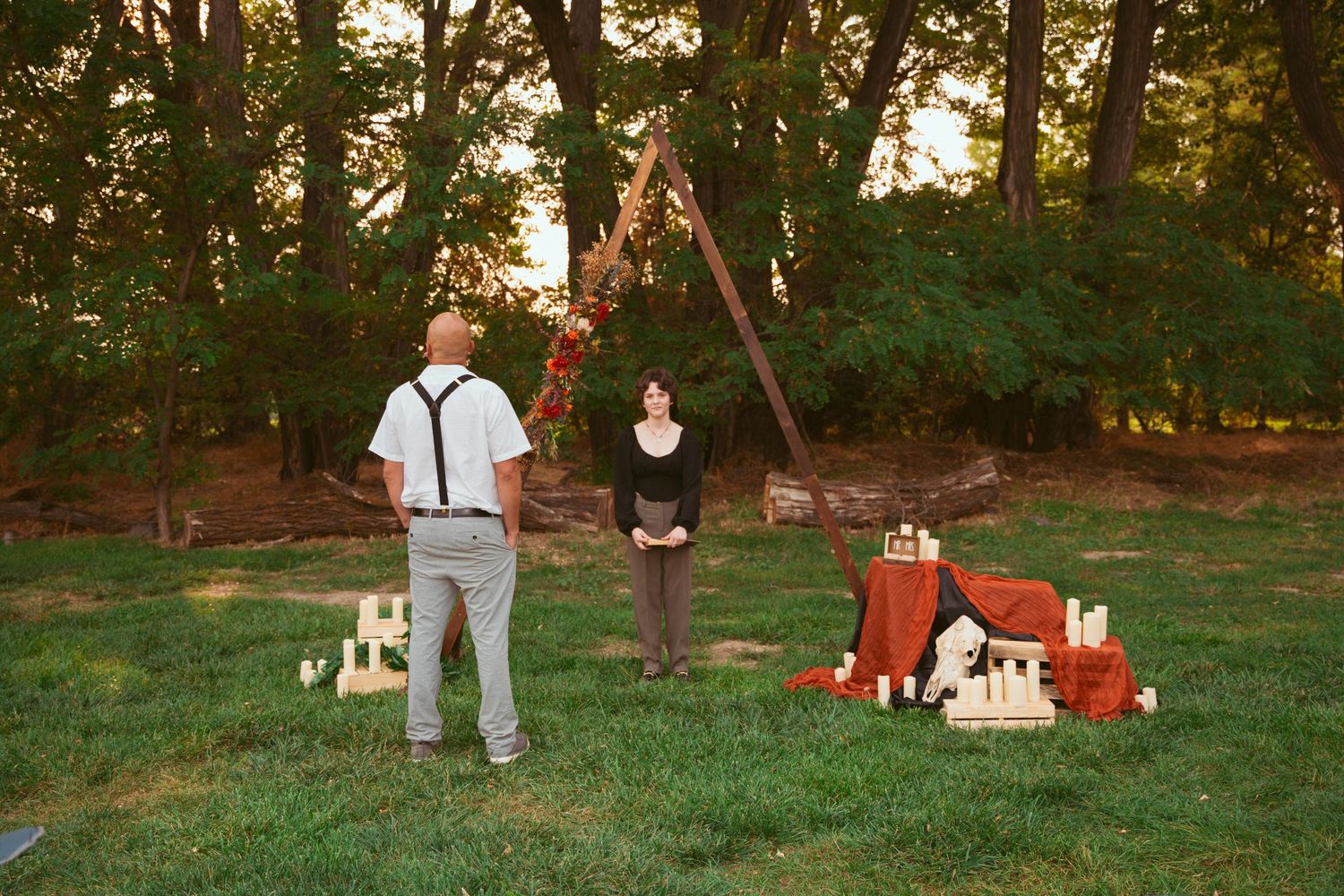 Wedding ceremony setup in a wooded area with guests seated on black chairs at sunset.