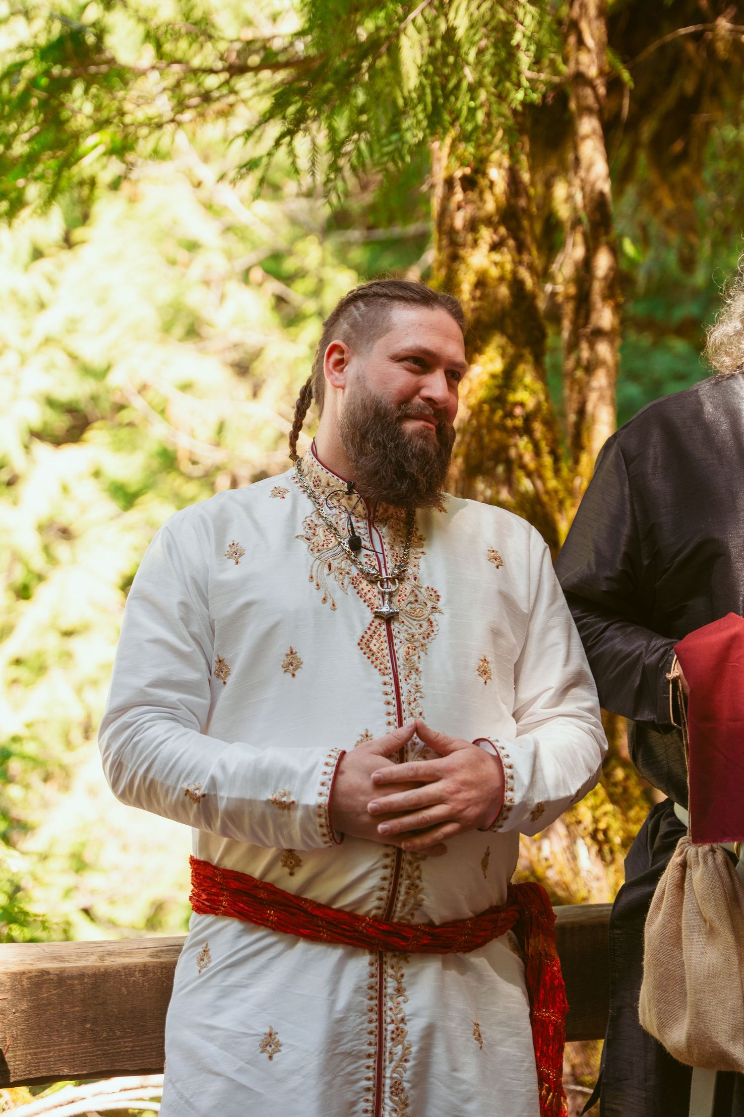 Medieval-themed wedding officiant in white embroidered garments gestures during forest ceremony.
