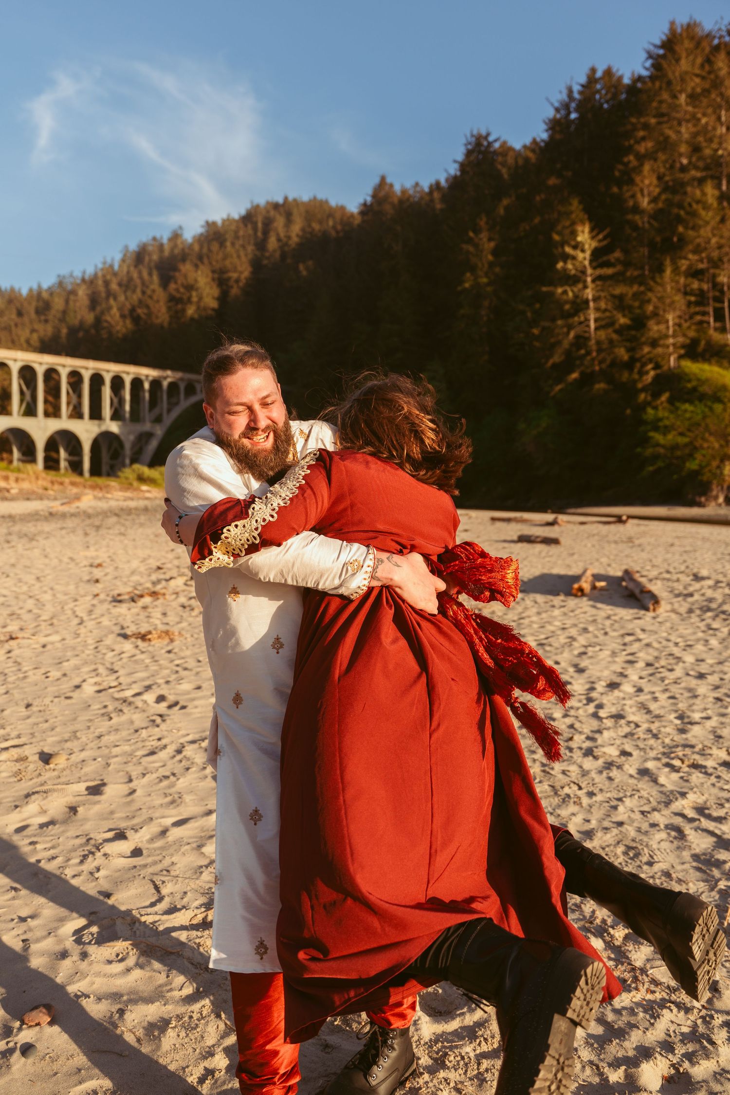 Two people in coordinated red and white outfits share a warm embrace on a sandy beach at sunset.