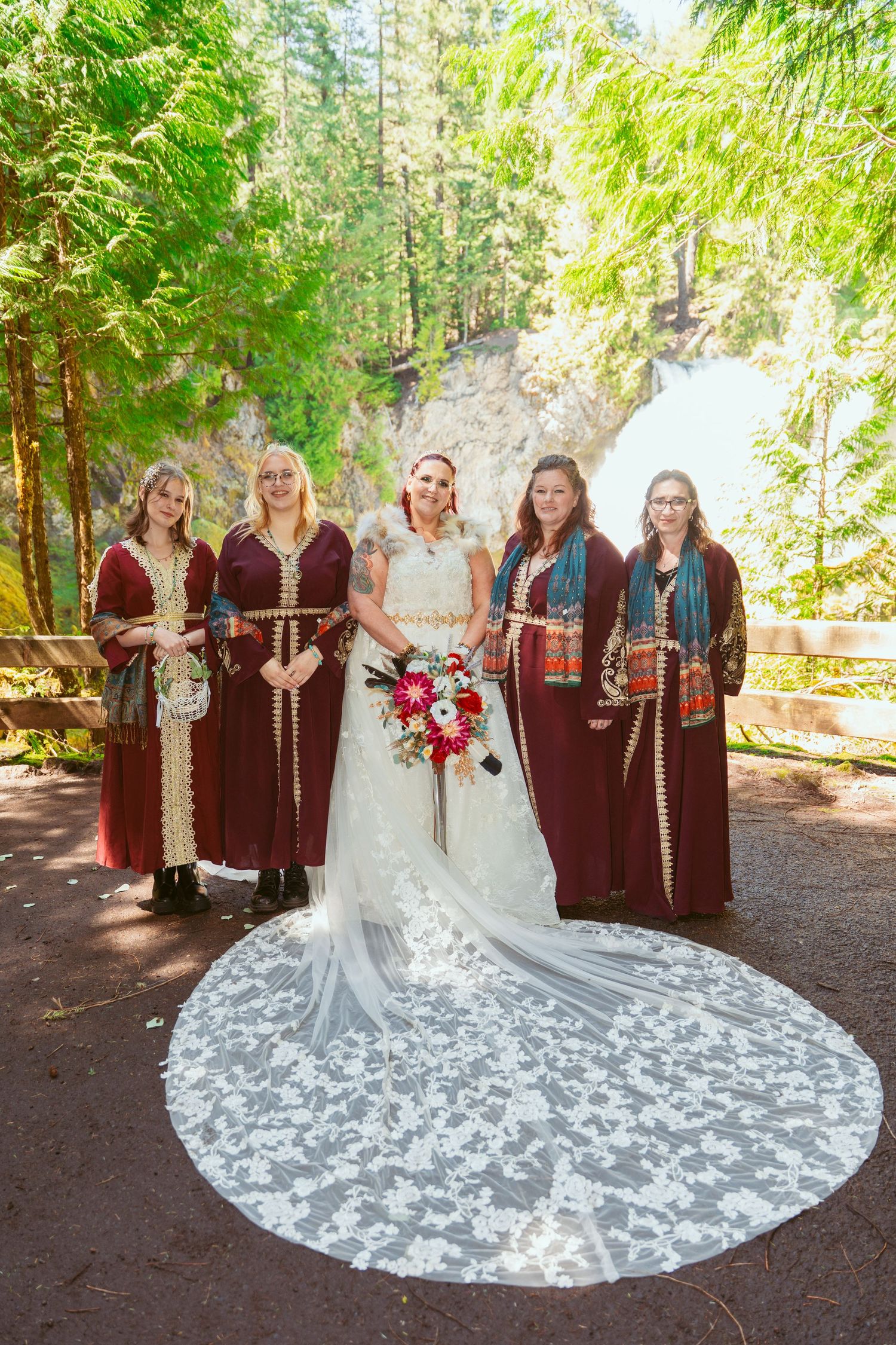 A wedding party in medieval-style attire poses in a forest setting with a stunning white lace dress train.