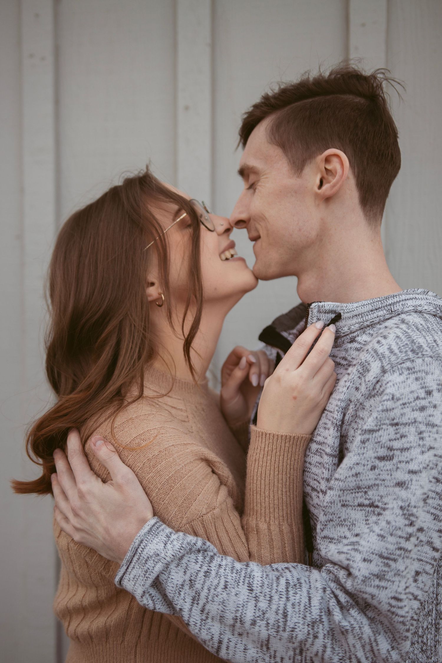 A romantic couple in cozy sweaters share an intimate moment while embracing against a white wall.