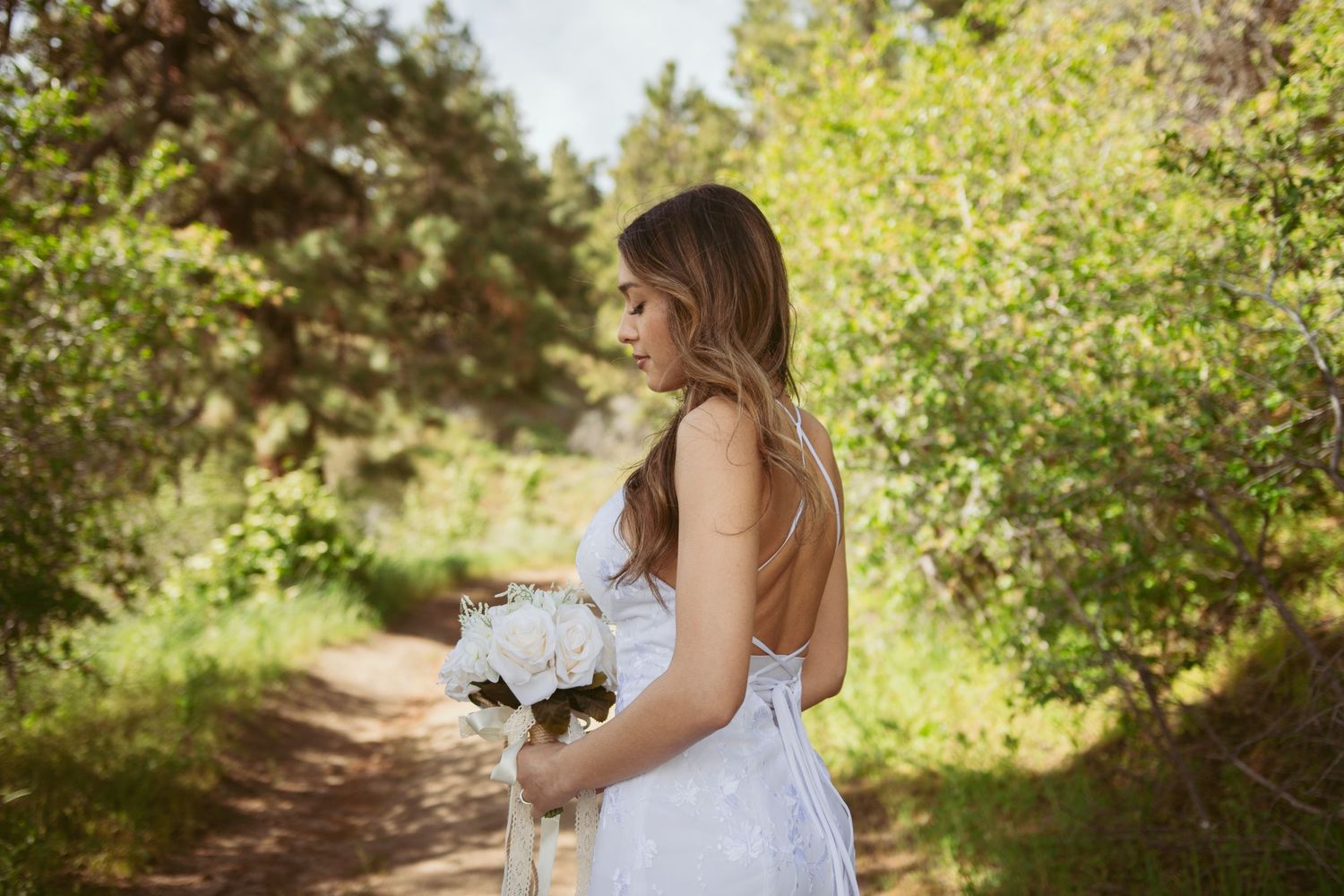 Bride in lace gown holds white bouquet while standing on forest path.